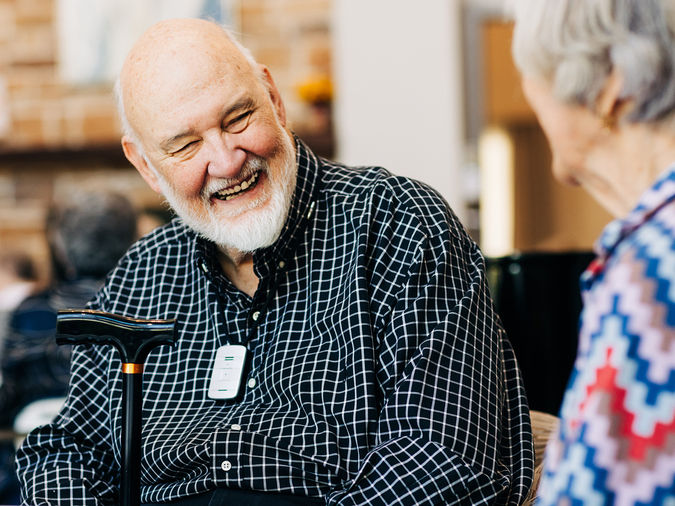 Senior man with a cane smiling warmly at an elderly woman in conversation, indoors.