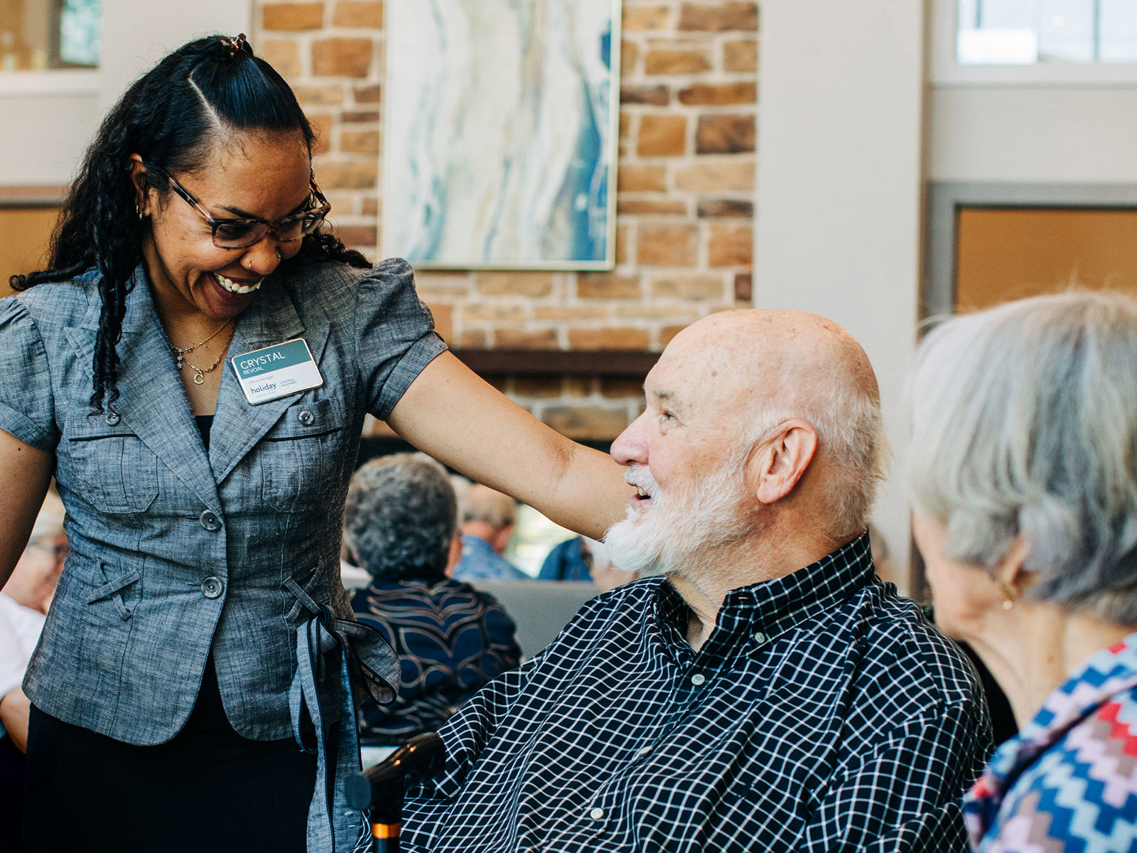 A smiling woman with glasses greets a senior man in a wheelchair, while another older person sits nearby in a cozy, well-lit room.