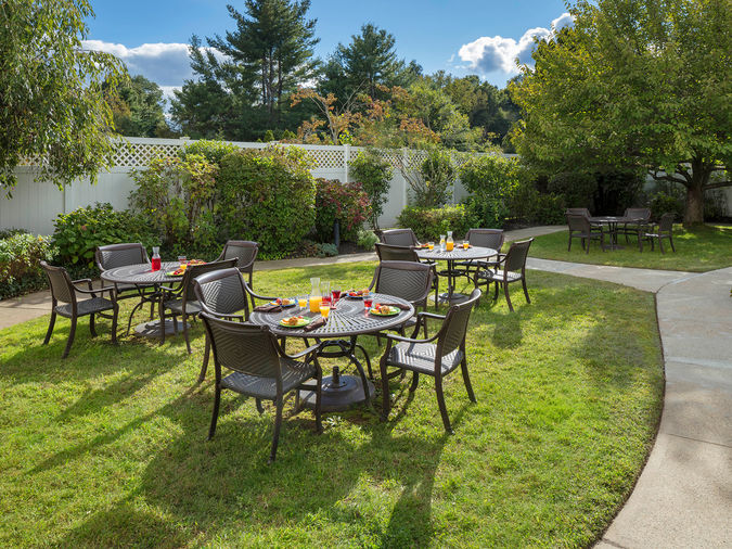 Outdoor patio with round tables set for dining, surrounded by grass and trees, under a partly cloudy sky.