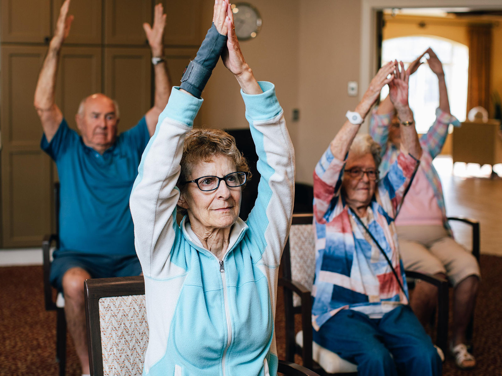 Seniors seated in chairs, participating in a group exercise, raising their arms overhead in a bright room.