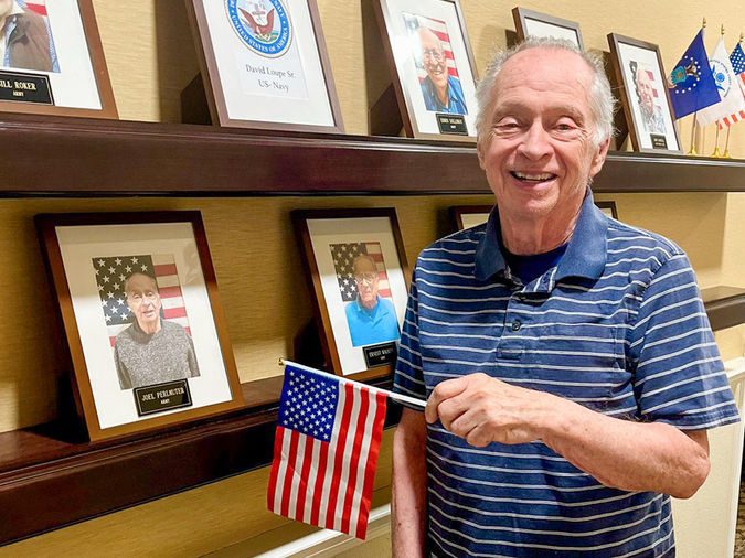 Older man smiling, holding a small American flag, stands in front of a wall displaying framed photos of veterans.