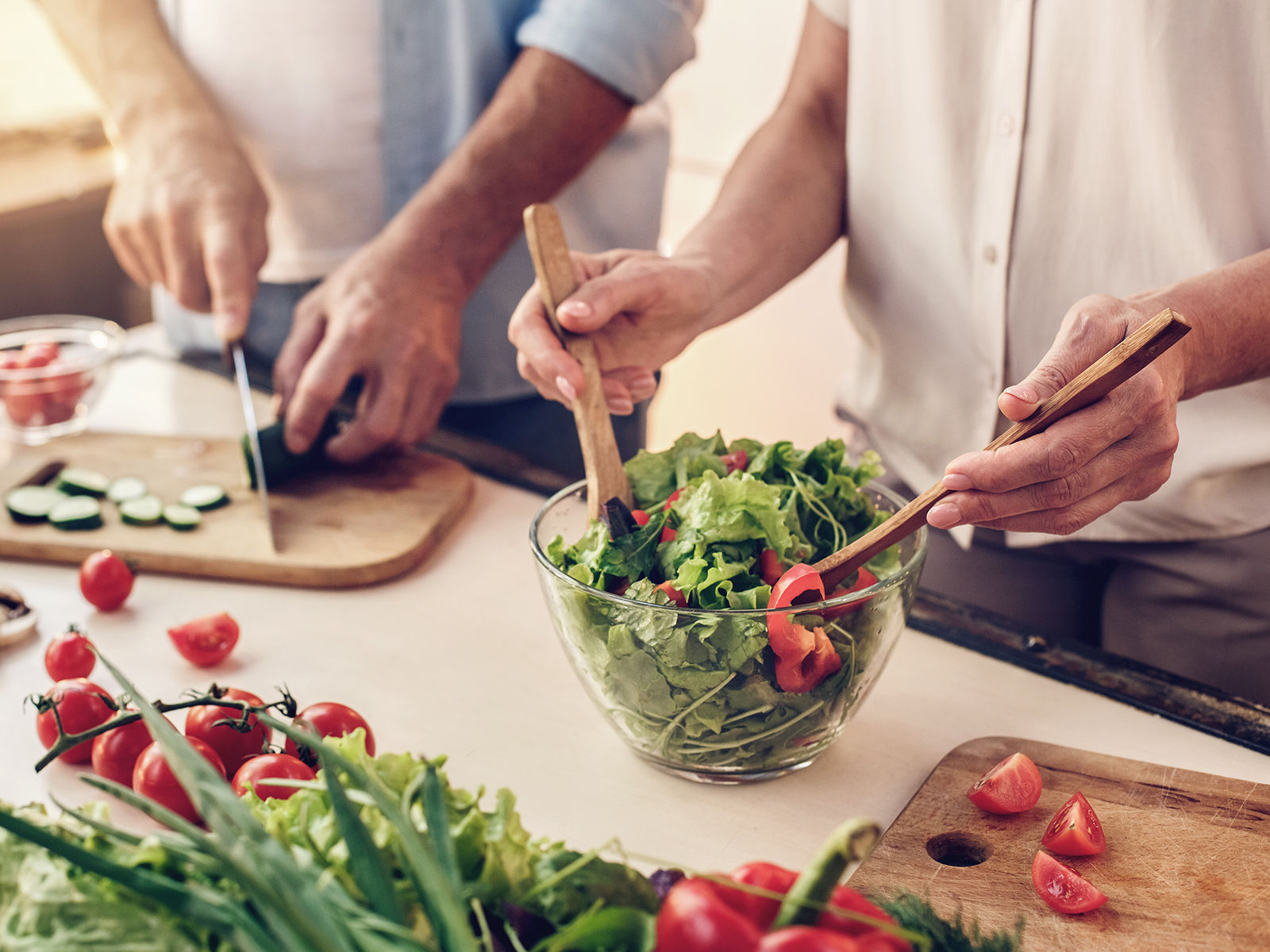Two people preparing a fresh salad with lettuce, tomatoes, and cucumbers on a kitchen counter with wooden utensils and cutting boards.