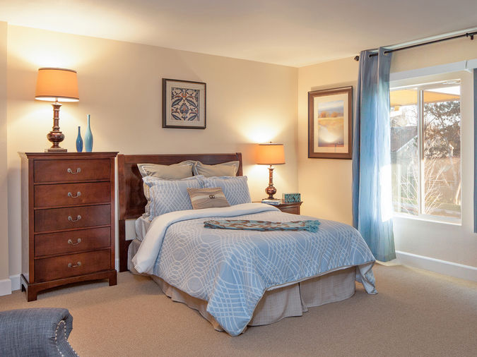 Cozy bedroom with a neatly made bed, wooden dresser, lamps, framed art, and a window with blue curtains letting in natural light.