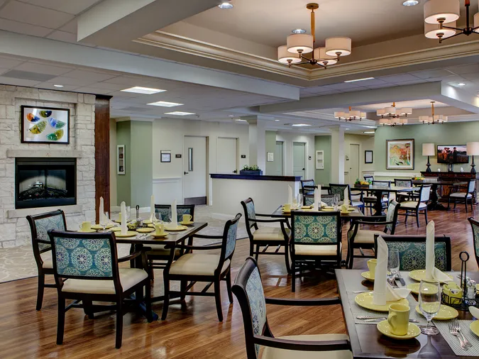Elegant dining area with neatly set tables, cushioned chairs, and a fireplace. Soft lighting and artwork create a welcoming atmosphere.