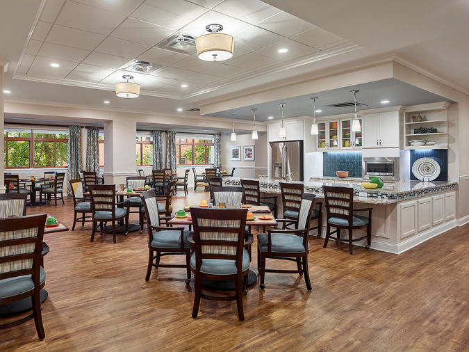 A spacious, well-lit dining area with wooden floors, neatly arranged tables and chairs, and an open kitchen with modern decor and lighting.