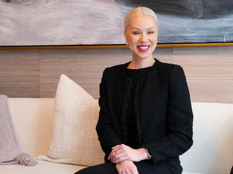 A woman with blonde hair in a black outfit sits on a white couch, smiling. A large abstract painting hangs on the wall behind her.
