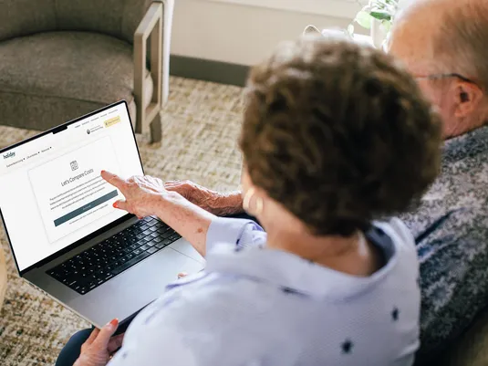 Older couple using a laptop, viewing a webpage titled "Let's Compare Costs" in a cozy home setting.