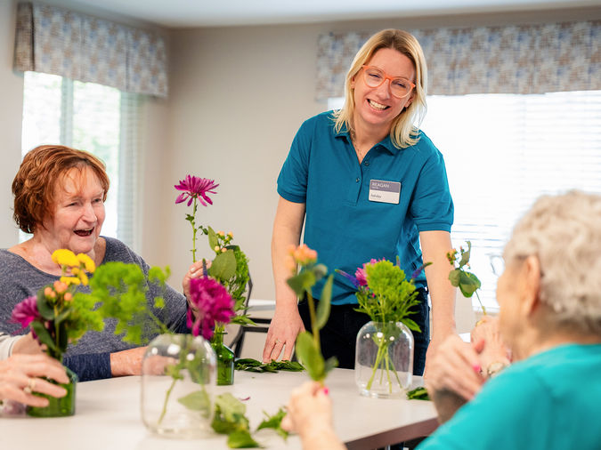 A smiling caregiver assists three senior women arranging flowers in vases at a bright, cheerful room with large windows.