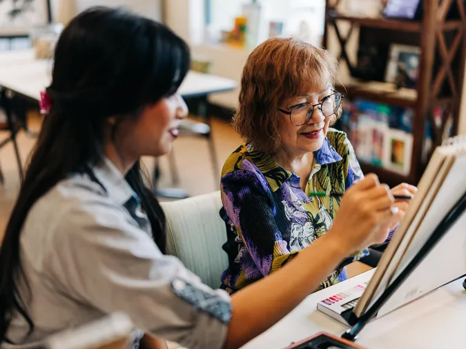 Older woman with glasses smiling as a younger woman guides her painting at an easel in a bright art studio.