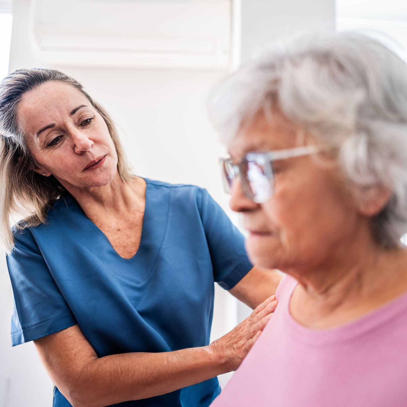 A healthcare worker in blue scrubs assists an older adult woman wearing glasses and a pink shirt in a bright room.