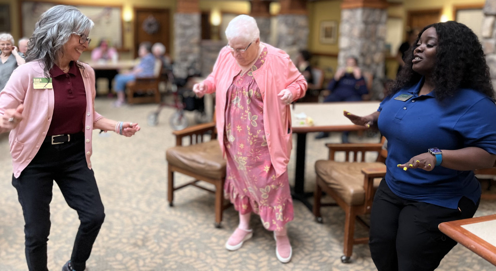 Three women joyfully dancing together in a room, with others seated in the background. They are smiling and appear to be enjoying the moment.