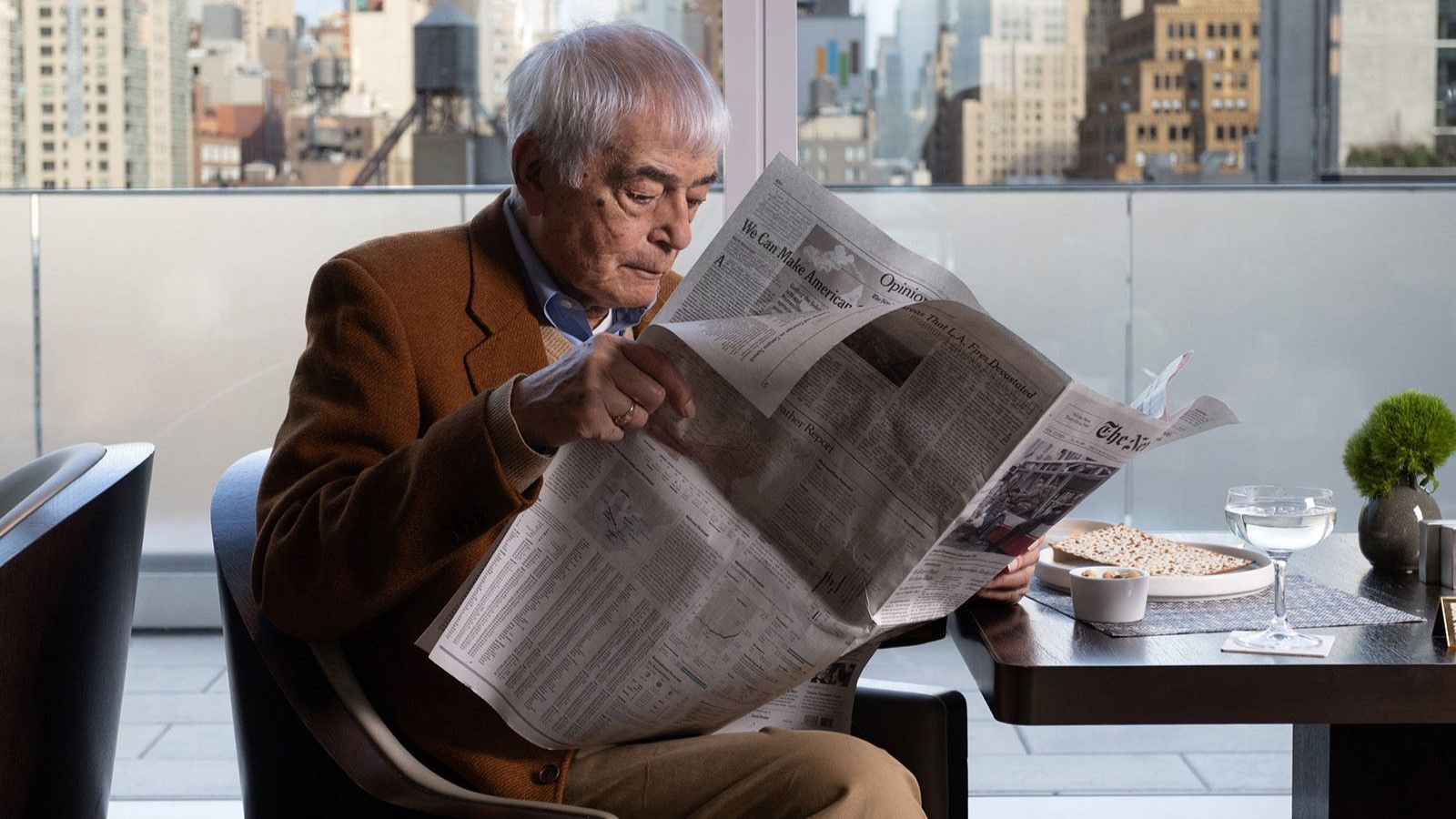 An older man sits alone at a restaurant table by a large window overlooking a city skyline. He is reading a newspaper while a plate of food, a glass of water, and a small vase with greenery sit in front of him. A ‘Reserved’ sign is placed on the table, and the bright daylight highlights the tall buildings outside.