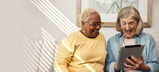 A black senior woman and a white senior woman laughing while holding a tablet