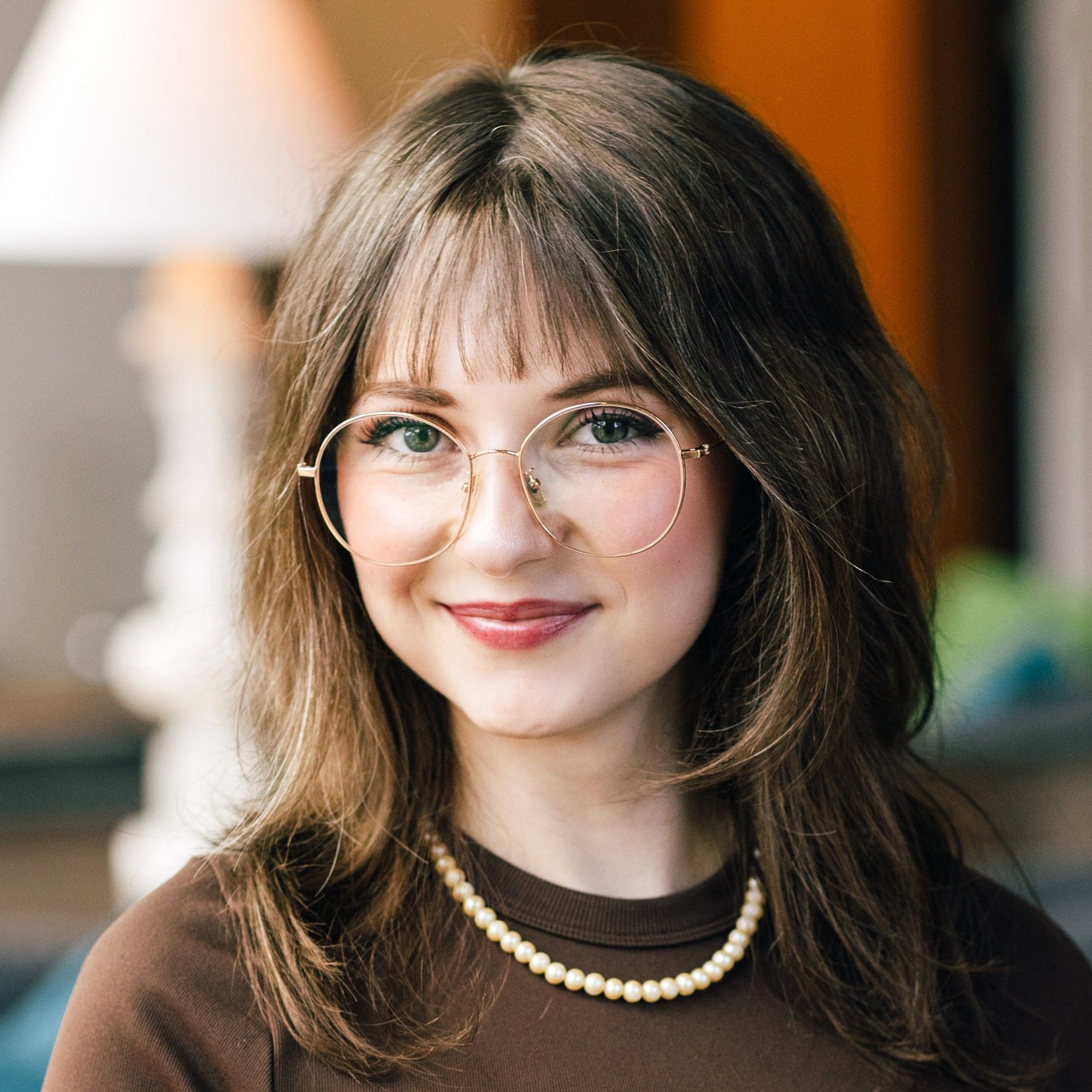 Smiling woman with wavy brown hair, large round glasses, wearing a pearl necklace and a brown top, indoors with a blurred background.