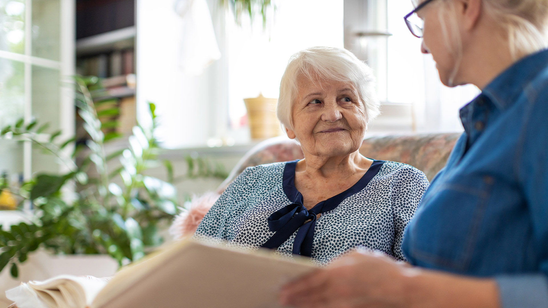 Senior woman and her adult daughter looking at photo album together on couch in living room