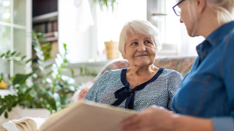 Senior woman and her adult daughter looking at photo album together on couch in living room