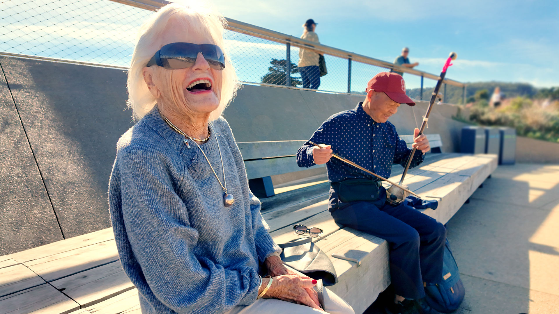 Older adult woman laughing on a bench while an older man beside her plays a traditional string instrument under a sunny sky.