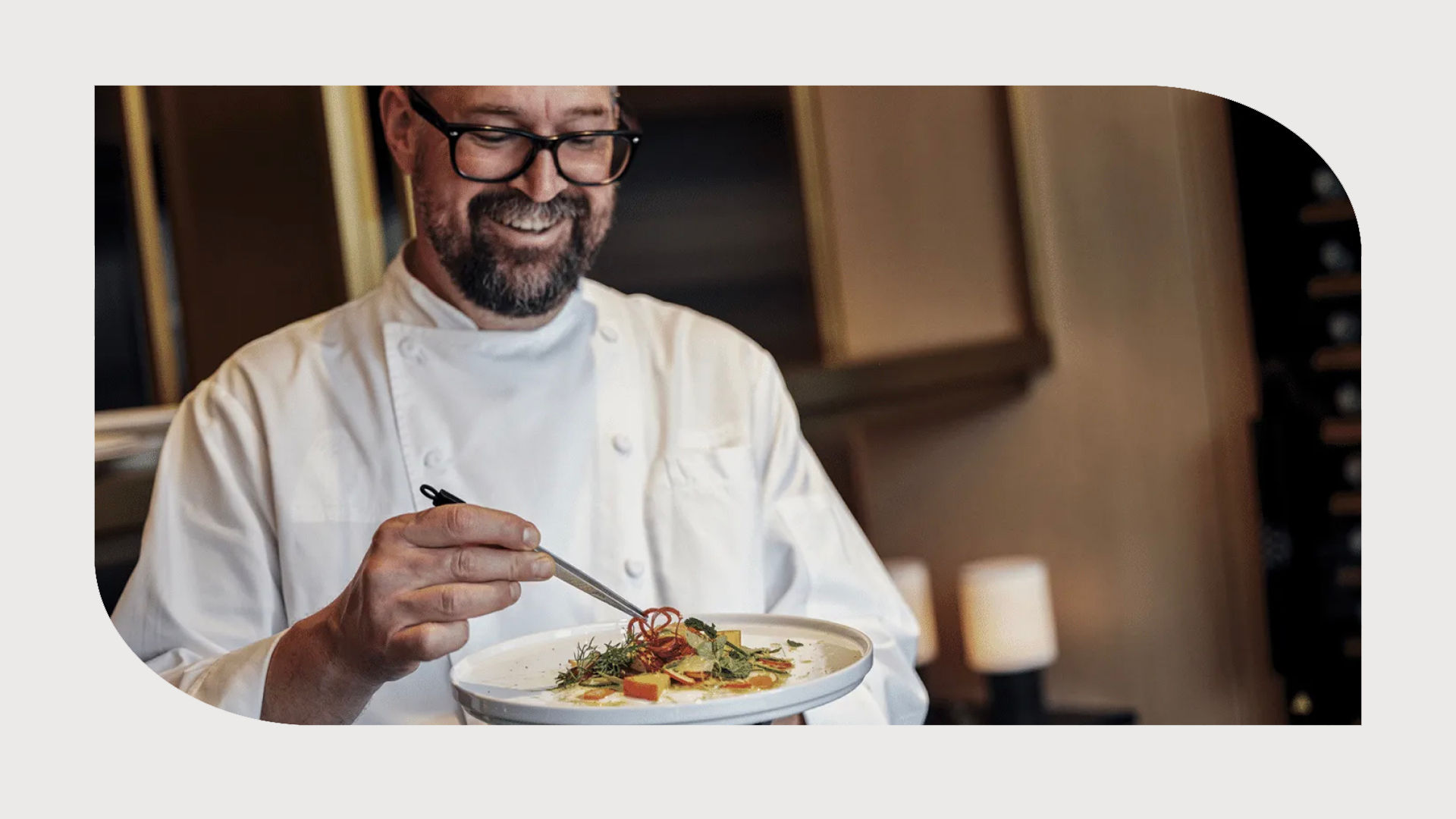 Chef in a white uniform smiling while garnishing a dish with precision in a warmly lit kitchen.