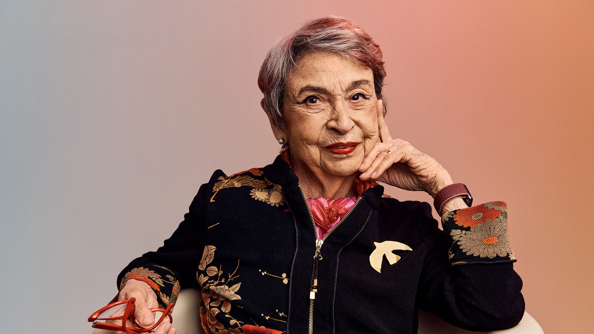 Older adult woman with short gray hair, wearing a floral-patterned jacket, holding red glasses, seated against a warm gradient background.