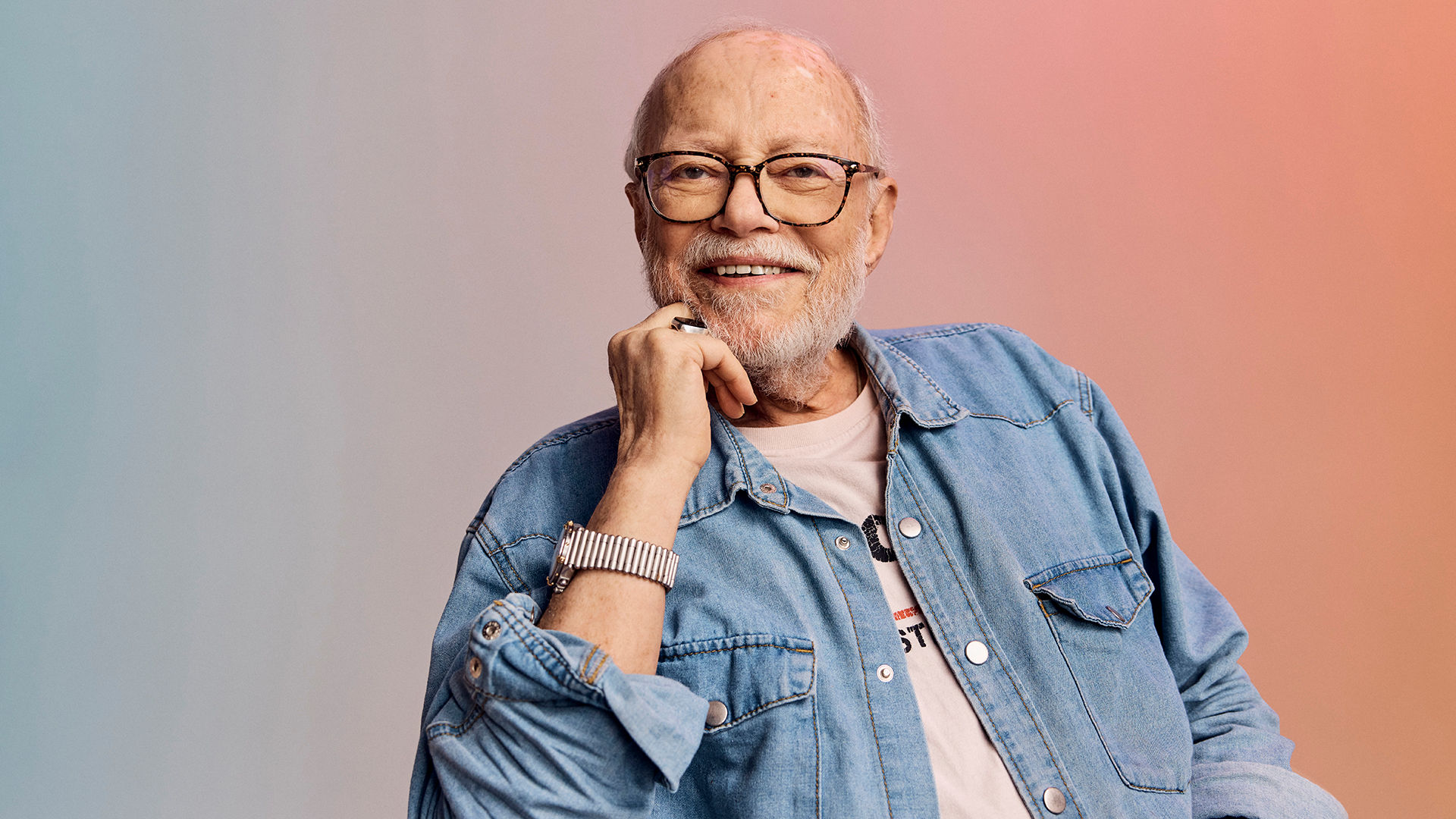 Smiling older man with glasses wearing a denim shirt, resting his chin on his hand against a soft gradient background.