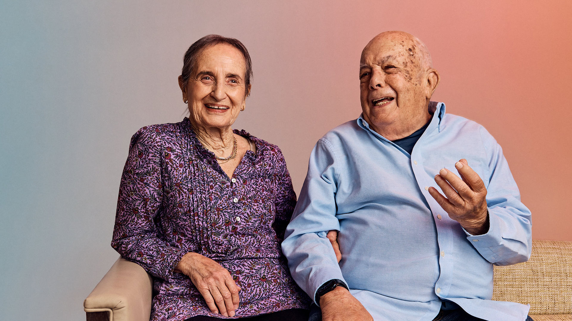 An older couple sitting together, the woman in a purple floral blouse smiles, while the man in a light blue shirt gestures animatedly.