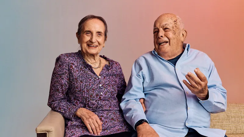 An older couple sitting together, the woman in a purple floral blouse smiles, while the man in a light blue shirt gestures animatedly.