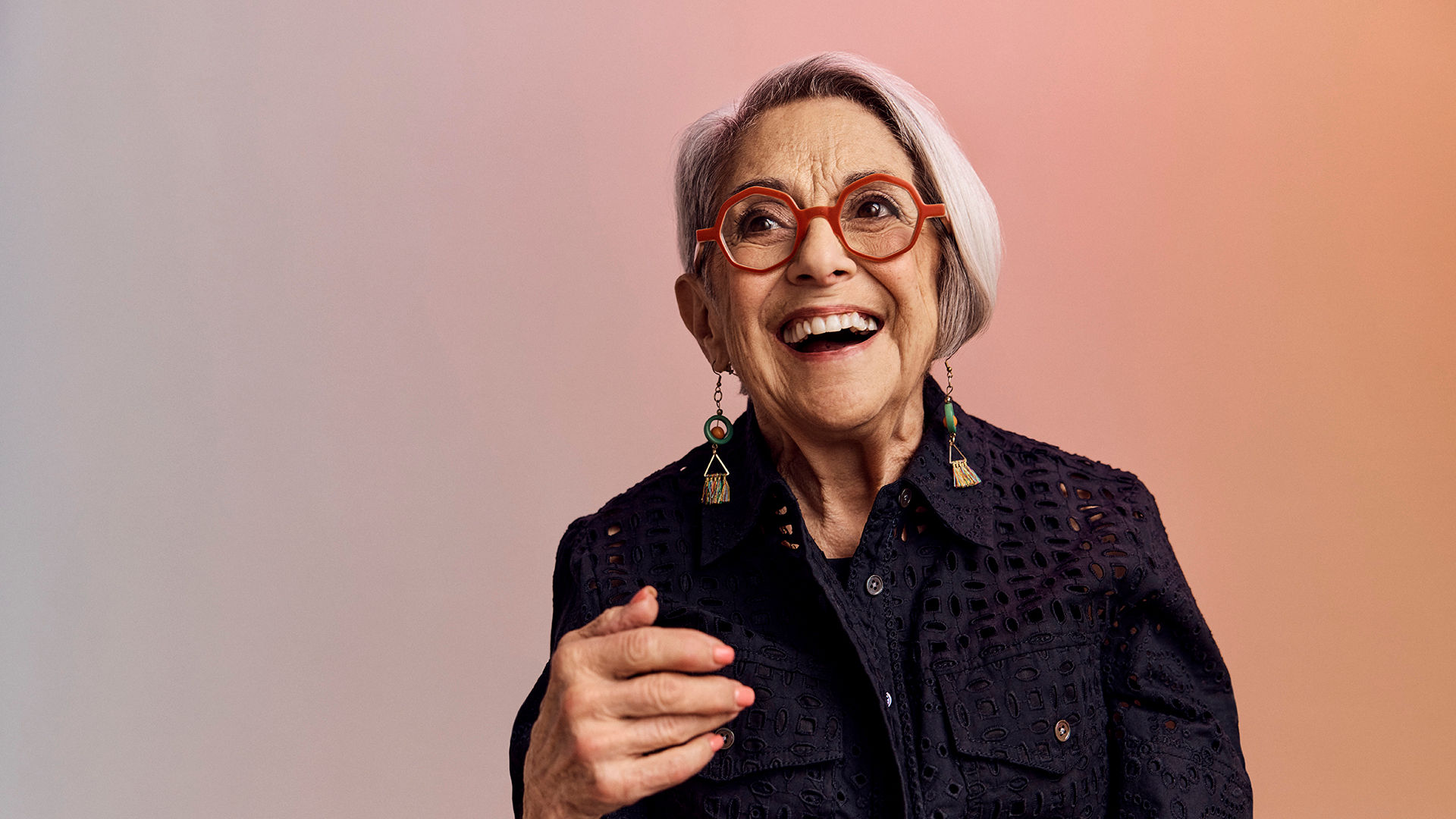Older adult woman with short gray hair, wearing red glasses, dangling earrings, and a dark lacy top, smiles joyfully against a soft gradient background.