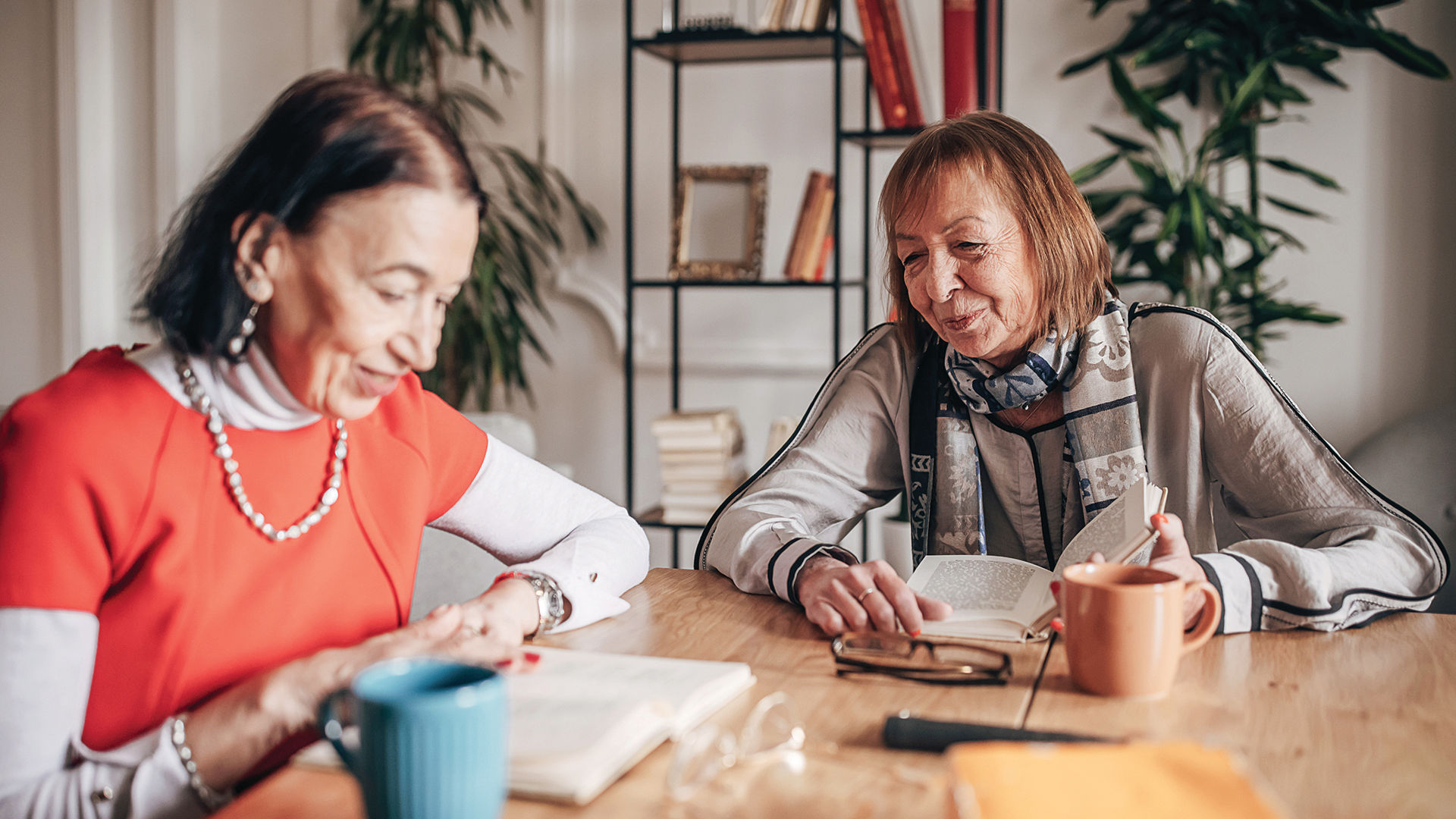 Two older women smiling and reading books at a wooden table with colorful mugs, surrounded by plants and a bookshelf in the background.