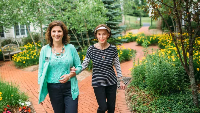 Two women smiling and walking on a brick path in a garden full of yellow flowers and lush greenery. One is assisting the other.