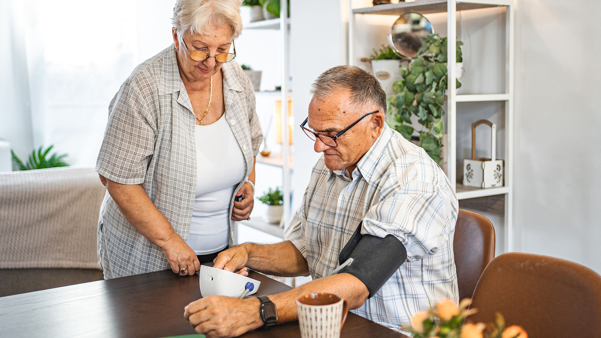 Man sitting at the kitchen table with a blood-pressure machine on his arm while his wife looks on