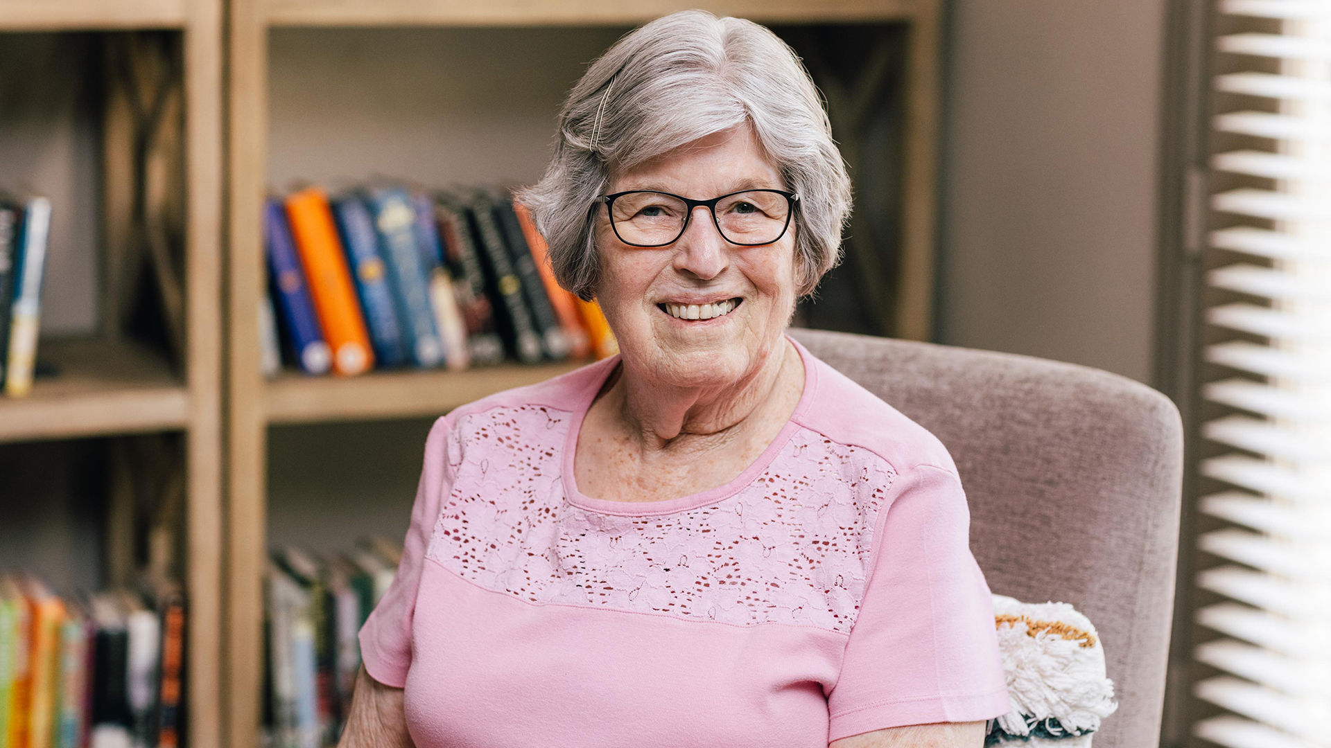 Holiday resident Glen, smiling with glasses and short gray hair, wearing a pink top, sits in front of a bookshelf filled with colorful books.
