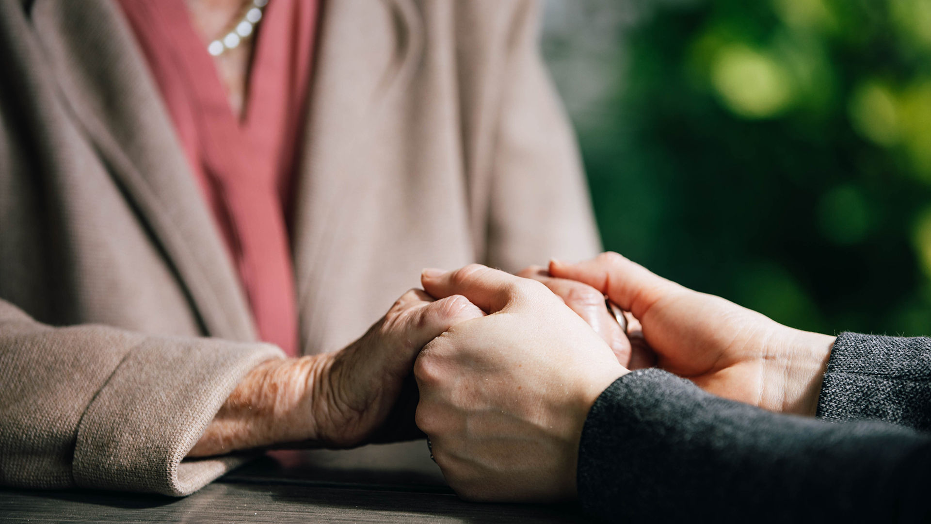 Two people sharing a comforting handhold across a table, one wearing a pink top and beige coat, the other in a dark jacket.