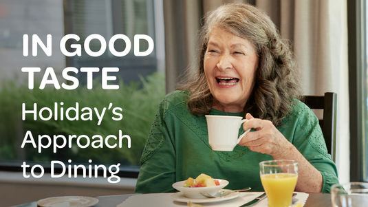 Senior woman in green top smiling while holding a cup at a dining table with breakfast, beside the text "In Good Taste: Holiday’s Approach to Dining."