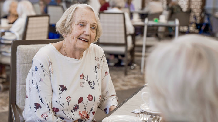 Older woman in a floral shirt smiling and conversing at a dining table in a communal setting.