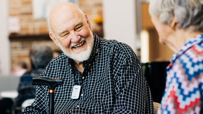 Older man with a beard smiling and holding a cane, conversing with a woman in a colorful blouse indoors.