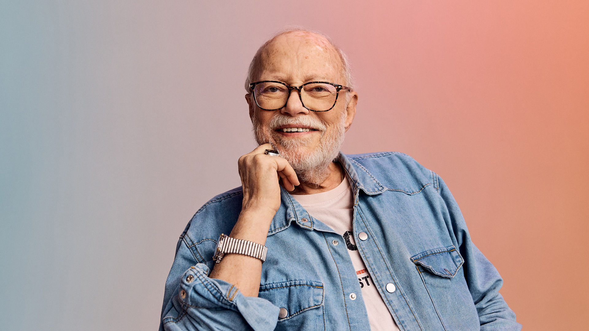 Smiling older man with glasses and beard, wearing a denim shirt and watch, against a soft gradient background.