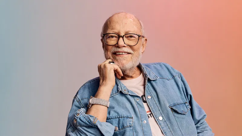 Smiling older man with glasses and beard, wearing a denim shirt and watch, against a soft gradient background.