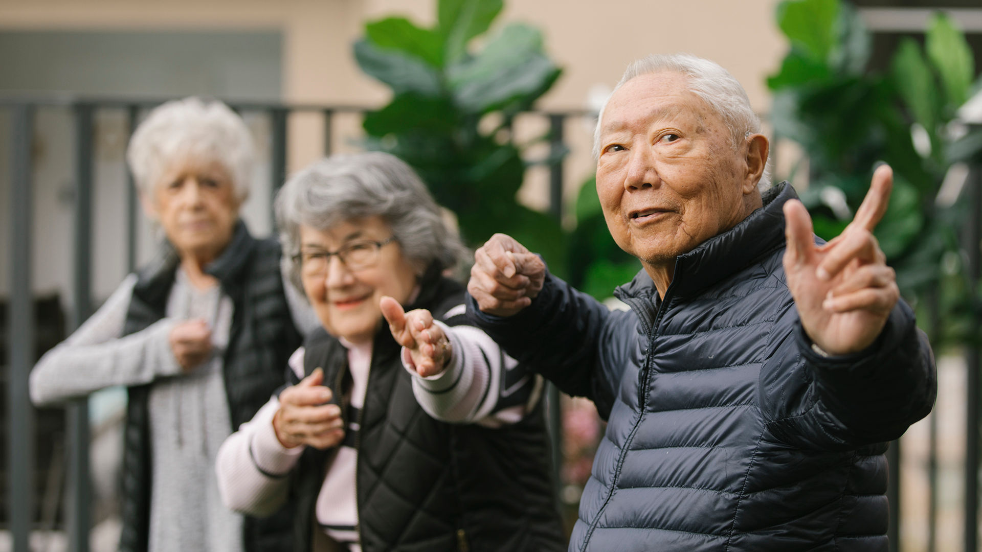 Three older adults in jackets, smiling and doing tai chi poses outdoors, with greenery in the background.