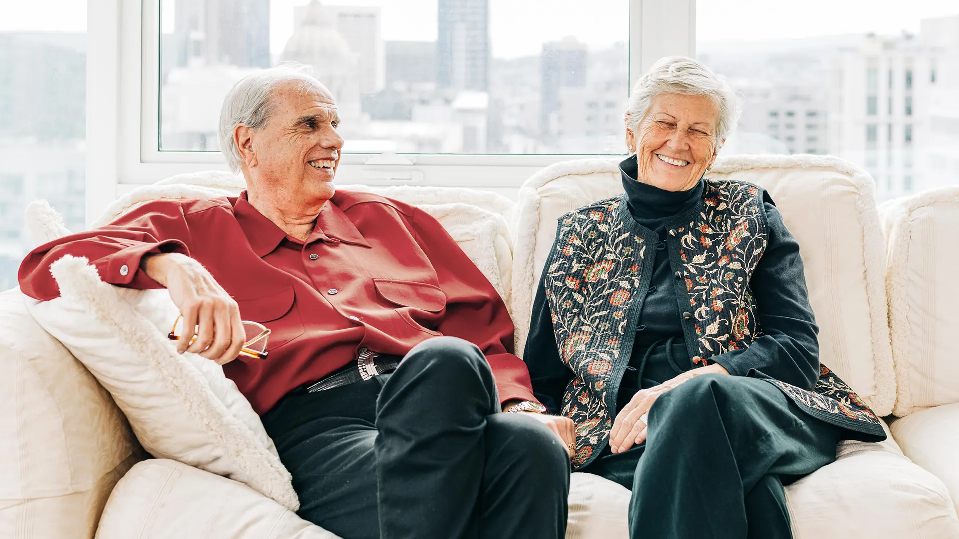 Older couple smiling while sitting on a cozy white sofa in a bright room with city views.