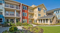 Colorful three-story apartment buildings with balconies, surrounded by green landscaping and a patio set, under a clear blue sky., click to stop animation