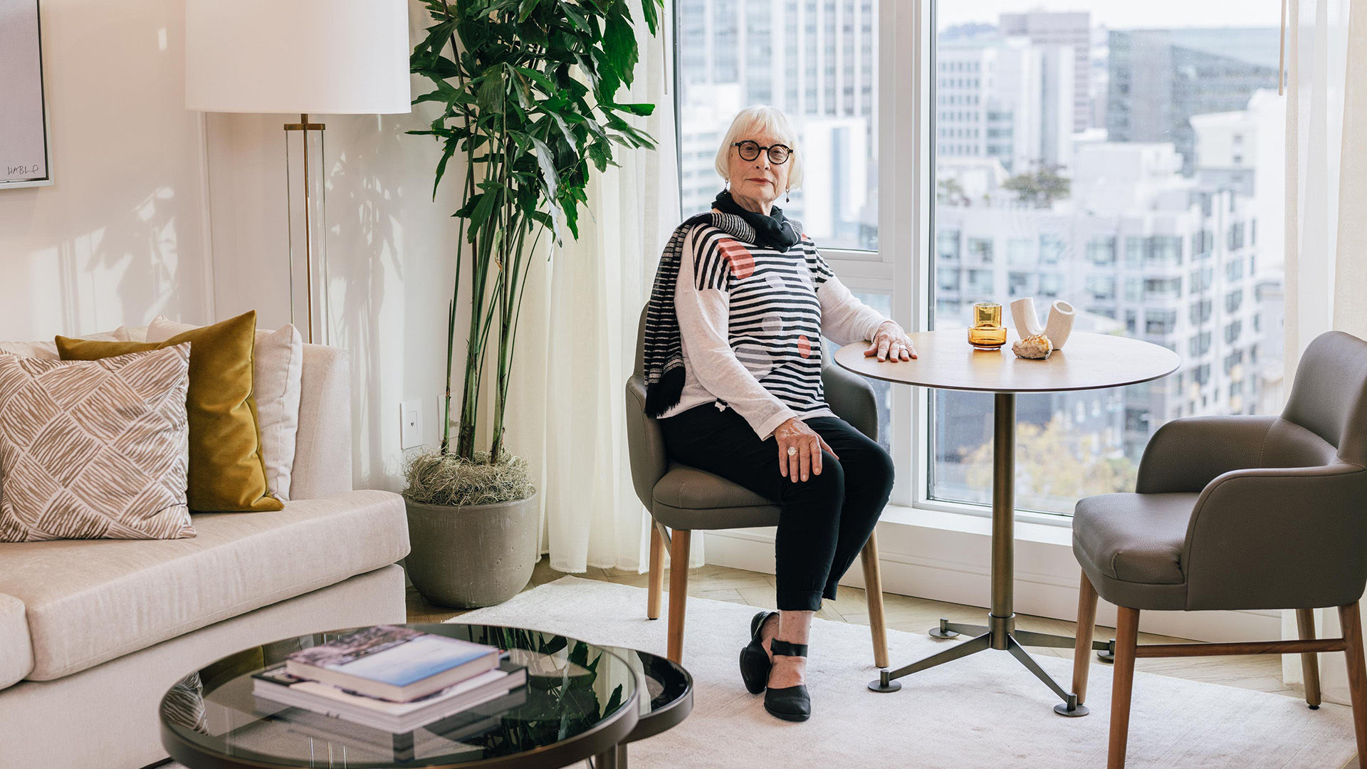 Coterie resident Carol sitting at a small table in a modern living room with large windows showing an urban view, and a sofa with cushions nearby.