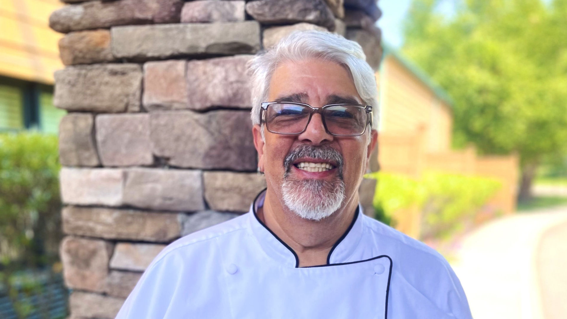 A smiling man in a chef's coat and sunglasses stands in front of a stone wall with trees in the background.