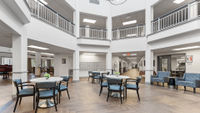 Spacious lobby area with round tables, blue chairs, and modern decor. Hallways extend on each side. Bright lighting and high ceiling., click to stop animation