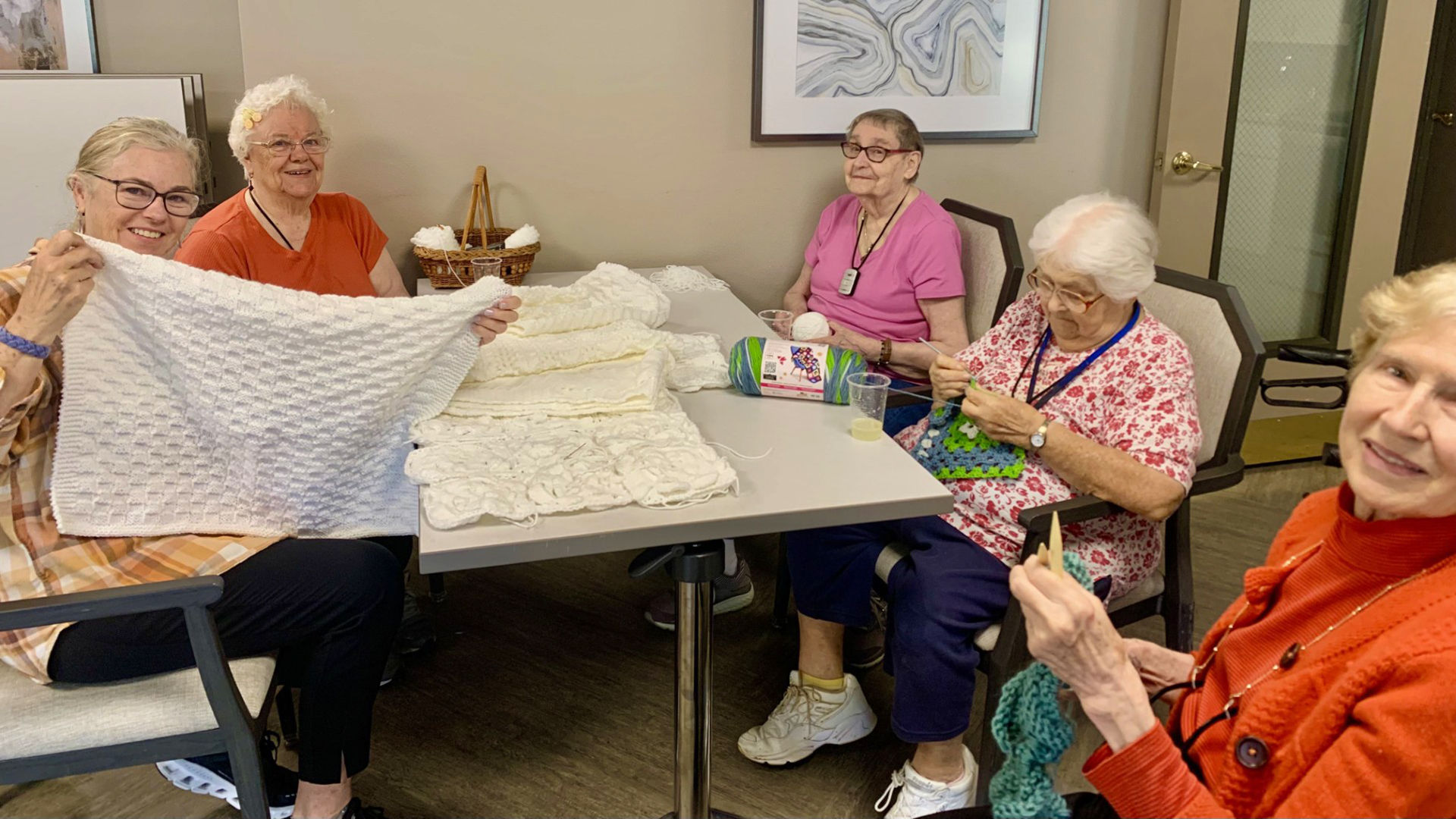 Five older adult women sitting around a table, knitting and displaying their knitted items, with yarn and baskets on the table.