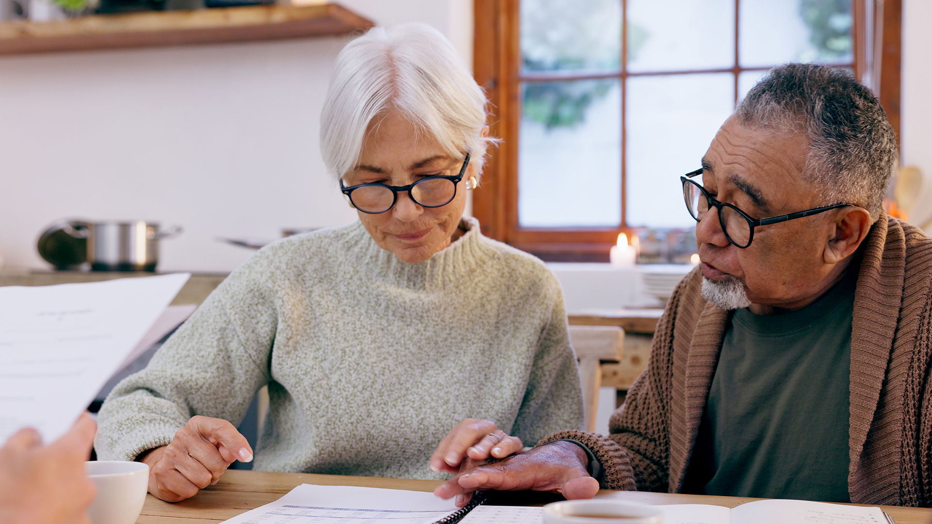 An older couple wearing glasses sits at a table, reviewing documents together while a candle burns in the background.