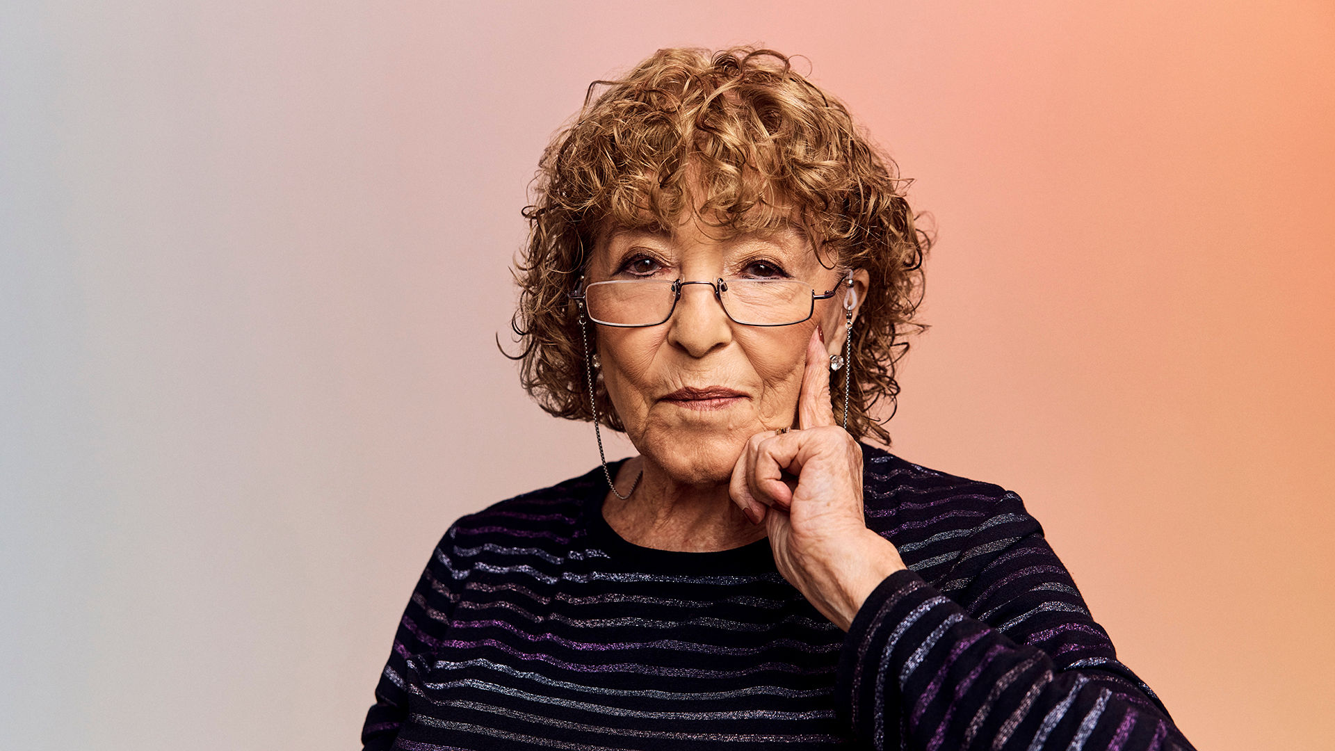 An older woman with curly hair and glasses, wearing a striped top, gestures thoughtfully with her hand on her chin against a soft gradient background.