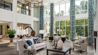 Sunlit two-story living room with floor-to-ceiling windows, blue patterned curtains, neutral sofas, wooden coffee table, and brass chandelier., click to stop animation