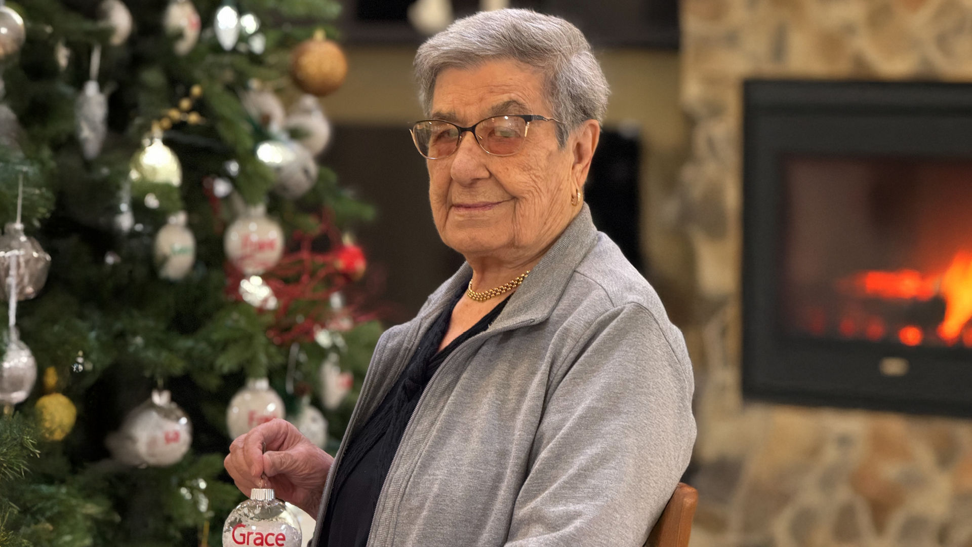 Senior person sitting by a Christmas tree, holding an ornament, with a fireplace in the background.