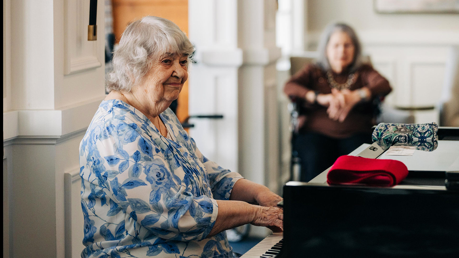 Older adult woman with gray hair playing the piano in a floral blouse, while another woman in the background sits and watches.