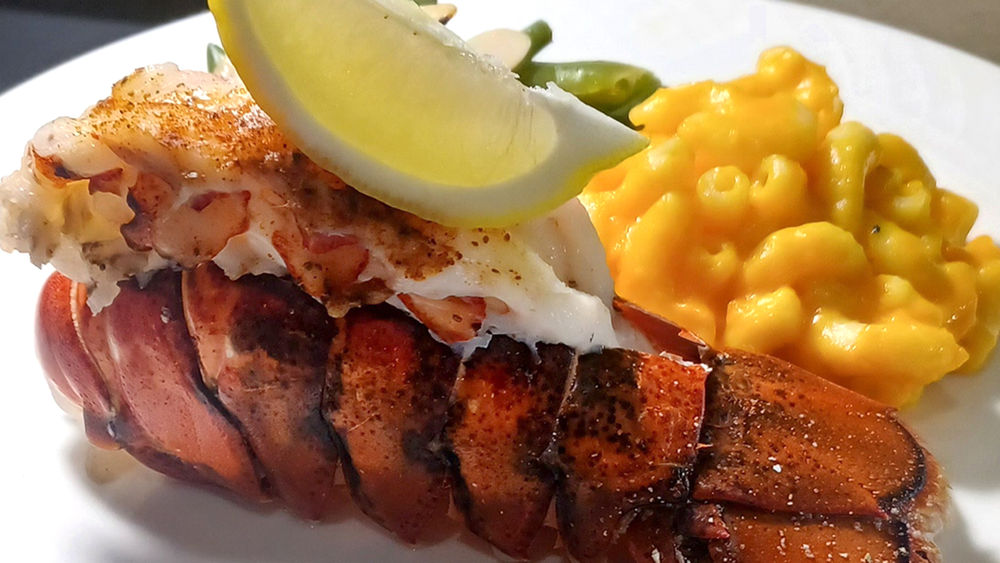 Lobster tail topped with a lemon wedge, served with mac and cheese and green beans on a white plate.